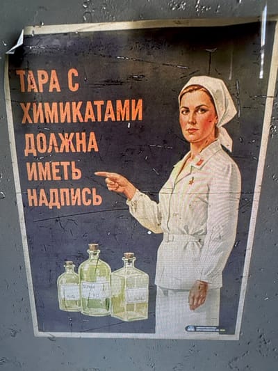 The image displays a photograph of a vintage Soviet-era safety poster. The central figure is a young woman, likely a laboratory worker or chemist, shown from the waist up. She wears a white lab coat or smock, a white head covering, and a red emblem on her collar. Her expression is serious and direct, as she gazes towards the viewer. With her right index finger, she points downwards towards a stack of laboratory bottles and the accompanying instructional text.

Three glass bottles, containing a light greenish or yellowish liquid, are depicted diagonally on the right side of the poster. Each bottle has a white label; visible text includes "HCL" (hydrochloric acid), "БЕНЗИН" (Benzin/Gasoline), and faintly "H2S" (hydrogen sulfide) on the uppermost bottle.

The poster features a dark blue or black background. The main safety message is presented in bold orange Cyrillic letters, oriented vertically along the left side. It reads: "ТАРА С ХИМИКАТАМИ ДОЛЖНА ИМЕТЬ НАДПИСЬ," which translates to "CONTAINERS WITH CHEMICALS MUST HAVE A LABEL."

The poster's aesthetic is characteristic of Soviet instructional art from the mid-20th century, emphasizing a clear, vital message. The overall tone is one of seriousness, highlighting the importance of chemical safety protocols. The photograph of the poster shows signs of age, including a slightly textured surface and minor wear.