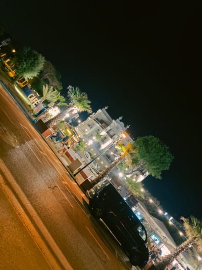 This is a night time, eye-level, oblique shot of a street scene in Rimini, Italy. The shot captures a building with several floors, featuring decorative architectural elements and illuminated windows. In front of the building, there is a cafe or restaurant area with outdoor seating, palm trees, and string lights. A black car is parked on the right side of the street. The street itself is paved and illuminated by streetlights, casting a warm glow on the wet surface. Several people are visible, some walking, others seated at tables, appearing to be patrons of the establishment. A large sign with the word "CAFE" in white block letters is visible on the left side of the frame. The sky is dark and clear, indicating it is nighttime.