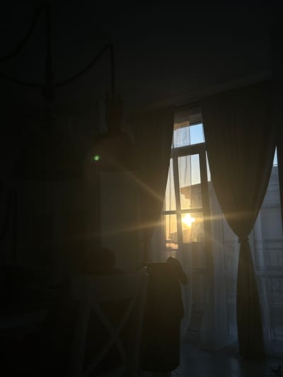 The image depicts an indoor scene at dusk or dawn, with sunlight streaming through a window. The window is partially covered by sheer, cream-colored curtains. The sun's rays create lens flare and cast strong light across the room, illuminating dust particles in the air. In the foreground, a dark silhouette of a person can be seen sitting in a white chair, facing away from the camera. To the right, another dark silhouette of a person is standing and appears to be looking out the window. The room is dimly lit overall, with shadows obscuring much of the detail in the background and to the left. The architectural style suggests a residential setting. The location within Chernomorets, Bulgaria, is not discernible from the visual information provided.