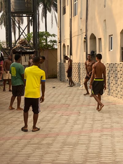 In Eziobodo, Nigeria, an outdoor scene captures seven young adult males gathered on a light-colored paved surface, featuring rectangular pavers interspersed with red lines. The setting is an alleyway or courtyard between a light-toned, multi-story building with arched ground-floor features and a brown and white brick base on the right, and a metal water tower structure with a dark water tank on the left. Several palm trees and green foliage are visible against a bright sky in the background.
The individuals are positioned across the paved area. One male, facing away from the viewer in the immediate foreground, wears a yellow short-sleeved shirt with blue trim, black shorts with white stripes, and dark flip-flops. To his left, another male wears a green t-shirt and dark shorts, standing barefoot. Further left, a shirtless male in light shorts is partially visible. Behind them, another shirtless male is seen smiling. On the right side of the frame, a shirtless male in dark shorts is smiling and facing left, displaying visible perspiration. Another shirtless male in dark shorts with a yellow-green stripe walks barefoot towards the right. Nearest the building wall on the right, a shirtless male in dark shorts stands, looking downwards, wearing dark flip-flops. The casual gathering suggests a relaxed interaction or pause in activity.