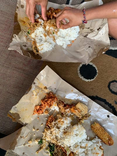Two servings of rice with side dishes are presented on brown paper wrappers, spread out on a woven mat with a red and black striped pattern. The upper serving shows hands in the process of eating. One hand, with a colorful braided bracelet on the wrist, is reaching into the food. The food consists of white rice topped with shredded meat, possibly chicken or beef, with chili, and some noodles. The lower serving displays rice alongside a portion of shredded meat with chili, and a piece of fried tempeh. The paper wrappers are crumpled and stained with food residue. The scene appears to be an outdoor dining setting in the city of Sukasada, Indonesia, possibly a roadside eatery or a picnic.