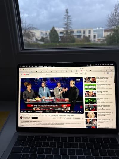 An indoor scene features a MacBook laptop placed by a window, showing an overcast sky and multi-story buildings with trees outside, indicative of a residential or urban environment in Reims, France. The laptop screen displays YouTube, with a video playing an ESPN MMA segment featuring four male commentators or analysts. On-screen subtitles read: "water. I'm just giving you a CHANCE TO DO THIS". The video's title is "Ian Machado Garry makes his case for title shot after beating Belal Muhammad | ESPN MMA", from the "ESPN MMA" channel with 2.47 million subscribers. The video is 5:10 into a 9:50 duration, has 149 thousand views from two days ago, and 2.9 thousand likes.
The YouTube page also suggests other videos, with titles visible including "Arman Tsarukyan DISMISSES Paddy Pimblett Title Hopes", "ALL OF ECONOMICS (No BS, No Fluff) in 20 minutes", "Why The UFC Sucks Now", "When Gaddafi Terrorized The World", "Technofeudalism: The Death of Capitalism and the Destruction of...", "CANCEL THIS FIGHT NOW!", and "Communism vs. Socialism". The Safari browser interface shows "youtube.com" in the address bar and "espn mma" typed into the search bar. The system clock in the menu bar indicates "Tue 25 Nov 4:55 PM". A portion of the laptop's keyboard is visible to the right.