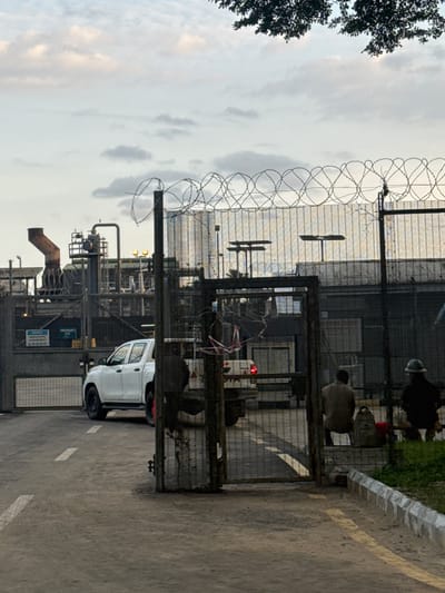 The image depicts an industrial facility in Igwuruta, Nigeria, secured by a tall metal fence topped with barbed wire. A white pickup truck is parked near the entrance gate. Industrial structures with pipes, tanks, and machinery are visible within the compound. Overhead, the sky is overcast. Some workers are visible within the compound, near the gate. The setting appears to be a roadside, with a curb and pavement markings visible.