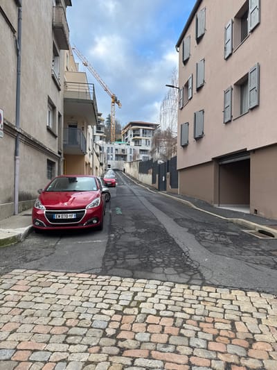 This image depicts an upward-sloping urban street in Le Puy-en-Velay, France, observed under an overcast sky. The foreground road surface consists of multi-toned cobblestones, transitioning to cracked and patched asphalt. A red Peugeot 208, bearing French license plate EW-070-YF, is parked on the left, partially on the raised curb. A second, darker vehicle is parked immediately behind it, followed by another red vehicle further uphill.
The left side of the street is defined by a multi-story light beige building featuring balconies with dark railings and multiple windows; a red circular "No Parking" sign with a diagonal line is affixed to a pole near this building. The right side features a multi-story light peach-colored building with a dark brown lower facade, containing multiple windows fitted with closed grey shutters and a dark, recessed garage entrance. A dark street light fixture extends from this building's facade.
In the background, the street ascends towards a hillside occupied by several modern multi-story buildings, some appearing to be under construction, characterized by concrete frames and multiple balconies. A tall yellow tower crane is prominently visible above these structures, indicating active development. Bare trees are dispersed across the hillside. A solitary, dark-clad individual is discernible walking uphill on the far left side of the road.