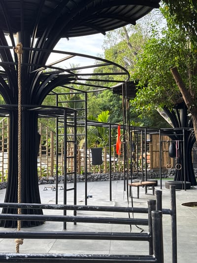 This image shows a multi-level outdoor gym facility in Baan Sri Thanu, Thailand. The structure is primarily constructed from black metal bars and features a combination of climbing frames, monkey bars, and rope elements. The ground is paved with concrete tiles. Lush greenery and trees surround the gym, blending the outdoor space with the natural environment. The perspective is from within the structure, looking outwards towards the surrounding landscape.