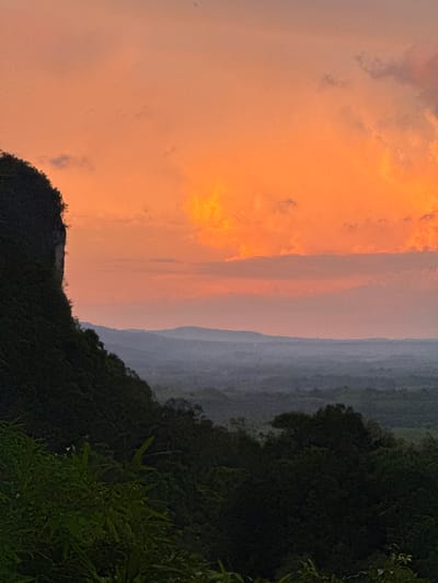 An outdoor, medium shot captures a dramatic sunset over a lush, mountainous landscape. The sky is ablaze with vibrant hues of orange and pink, with fluffy clouds scattered across the expanse. In the foreground, a steep, dark green mountainside covered in dense vegetation slopes downwards. The lower portion of the image features the shadowy canopy of a thick forest. In the distance, several layers of hazy, muted blue mountains recede into the horizon. The overall scene suggests a remote, natural setting, potentially a viewpoint overlooking a valley. The specific location is identified as Ban Khao Tang, Thailand. There are no people or discernible man-made structures within the frame.