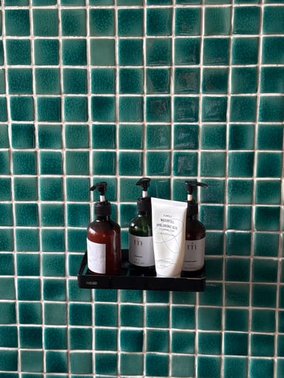 The image shows a black shelf attached to a wall covered in square, teal-colored tiles. On the shelf, from left to right, are three bottles and one tube of product. The first bottle is a dark brown glass bottle with a black pump dispenser. It has a white label with a pattern of vertical wavy lines. The second bottle is green glass with a black pump dispenser and a white label. The third bottle is also dark brown glass with a black pump dispenser and a white label. In front of the green bottle and to the right of the dark brown bottle is a white tube with a black cap. The tube has text that reads "JUMISO WATERFULL HYALURONIC ACID CLEANSING FOAM". The shelf has the word "HOBBY" printed on its front in white. The image is taken from a slightly elevated angle, looking down at the shelf and its contents against the tiled background. The location is South Quy Nhon Ward, Vietnam.
