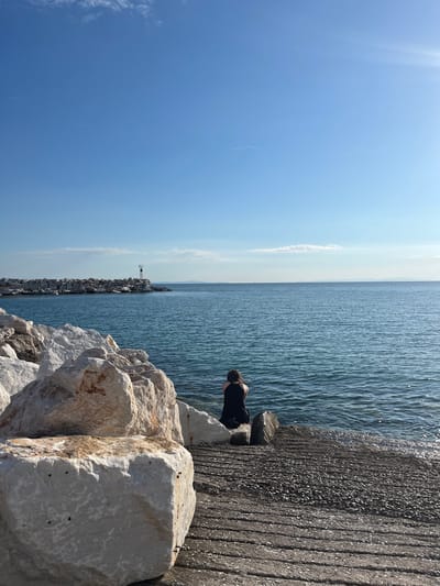 The image shows a person sitting on rocks by the sea in Potos, Greece. The person, wearing a black garment, faces the water. The sea is calm with gentle waves. In the distance, a breakwater made of large rocks extends into the water, with a small lighthouse on its end. The sky is clear blue with a few wispy clouds. Large, light-colored rocks are in the foreground, and a concrete pathway leads to the water's edge. The sun is shining, casting a bright light on the scene.