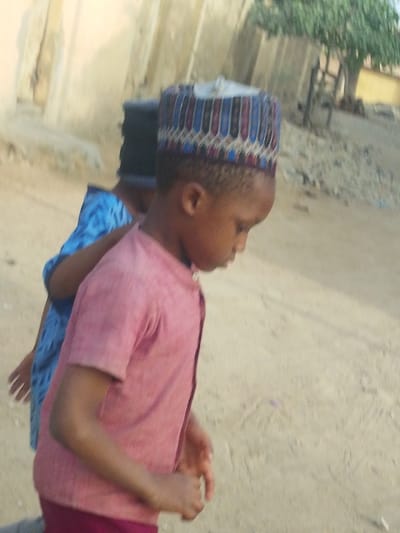 Two young individuals are captured outdoors on a light-colored sandy or dirt surface in Garun Gawa, Nigeria. In the foreground, a child with dark skin, wearing a pink short-sleeved collared shirt and a multi-patterned cap (kufi) with blue, red, black, and white elements, stands with their head bowed, looking downwards. Behind this child, partially obscured, is another individual wearing a blue patterned garment and a dark head covering. A light-colored, possibly concrete or mud-brick, building structure is visible in the left background, while a tree with green foliage is present in the upper right background. The scene is illuminated by natural light.