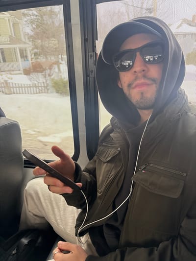A person wearing sunglasses and a hooded jacket sits inside a vehicle with a window showing an outdoor scene. They are holding a dark-colored device. The jacket has zipper pockets, and a white cable is visible near the neck area. The person has light skin, a beard, and their head is tilted to the side. Outside, a beach area is visible through the window.