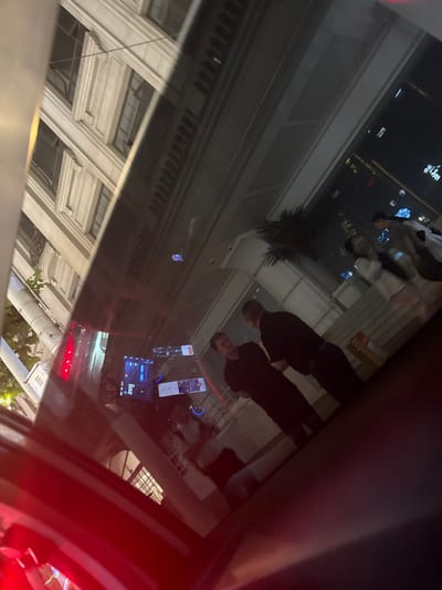 This media file captures a nighttime or late evening urban scene, viewed through a glass surface, likely a vehicle window. The image is dominated by reflections, revealing multiple layers of depth and activity.
**Subjects:** The most prominent subjects are two men reflected in the center-right of the image. Both wear dark, short-sleeved tops and have short hair. One man is in the foreground, facing slightly left, with his profile clearly visible. Behind him, another man’s face and upper body are also visible, similarly oriented. Further back and to the left in the reflection, other blurred figures, including one in a white shirt, suggest more people are present on the street or in an adjacent area. A large, dark, palm-like plant is also part of the reflection. On the far left and bottom-left, the ornate, classical-style facade of a building with columns, windows, and decorative cornices is strongly reflected.
**Objects:** Within the vehicle itself, on the right side of the image, two electronic screens are visible. The top one appears to be a smartphone showing an application interface. Below it, a car's infotainment screen displays various icons and text. A strong red light, possibly a vehicle's tail light or a traffic signal, is visible on the far right edge of the image, bleeding into the glass. The dark frame around the reflection suggests the interior of a car.
**Activity or event taking place:** The scene depicts an urban setting at night, with people standing or moving on a street or within a public building, as seen through a reflective surface. The presence of electronic screens and the car-like interior elements suggest the viewer is inside a vehicle, possibly waiting or traveling, and observing the illuminated reflections of the city.
**Notable details:** The time of day is clearly night or late evening due to the pervasive artificial lighting and dark ambient conditions. The architectural style of the reflected building, with its classical elements, provides a location cue consistent with areas in Shanghai, China, known for European-influenced architecture. The reflections are sharp, suggesting clear weather. No specific emotions are discernible from the reflected individuals.
**Visible text:** The electronic screens display text, predominantly in Chinese characters. On the car's infotainment screen, numbers like "25℃" and "4°C" are visible, likely indicating temperature.