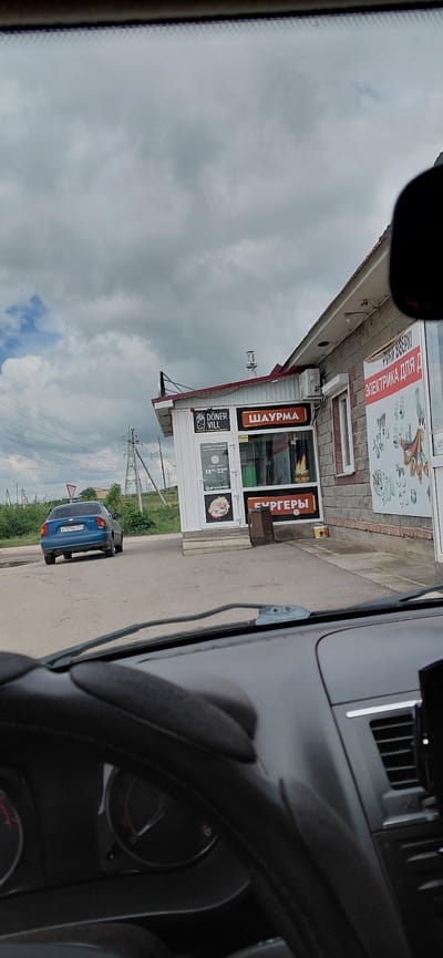 The image was taken from inside a car, looking out towards a roadside food establishment in Agapovka, Russia. The establishment appears to be a small building with a red roof, constructed with brick and white siding. Signs on the building indicate it sells "DÖNER VILL," "ШАУРМА" (Shaurma), and "БУРГЕРЫ" (Burgers). There is also a sign that reads "ЭЛЕКТРИКА ДЛЯ Д" (Electrics for D). The time displayed on one of the signs is 10:00-22:00, suggesting opening hours.
Outside the building, on the paved road, a blue Chevrolet sedan is parked. The car's license plate is partially visible and reads "X751KC774". The environment is somewhat overcast with a cloudy sky. Power lines and utility poles are visible in the background, along with some grassy areas and trees. A red triangular traffic sign is also visible on the left side of the road. The interior of the car, including the dashboard and steering wheel, is visible in the foreground.