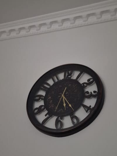 This media file contains a photograph of a dark-framed, analog wall clock with Roman numerals. The clock face has an ornate, dark design. The hour hand points to approximately the 2, and the minute hand points to approximately the 4. The clock is mounted on a light-colored wall. To the left of the clock, a decorative white molding runs vertically down the wall. The molding has a repeating geometric pattern. The overall setting appears to be indoors, likely a room in a home or office. There is no visible text in the image. The lighting suggests it is daytime, and the scene is static with no activity taking place.