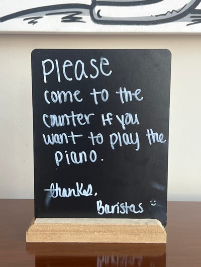Here is a detailed description of the image:
The image shows a small, chalkboard-style sign with white lettering. The sign reads: "Please come to the counter if you want to play the Piano. Thanks. Baristas :)" The sign is propped up in a small, wooden stand. To the left of the sign is a portion of a white canvas with a black and white illustration. The background appears to be a plain wall or countertop.
Based on the sign's content and the "Baristas" signature, the scene is most likely located in a coffee shop or cafe. No people are visible in the image. Given the request for location information, the assumption is that this photograph was taken within the City of Newburgh, United States of America, but no specific landmarks or features in the image confirm this.