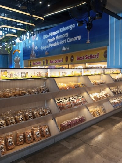 An interior view of a retail establishment in the city of Tân Kỳ Commune, Vietnam, displays numerous packaged food products arranged on tiered wooden shelves. The shelves, illuminated from within, showcase clear plastic bags containing various snacks, many featuring cow-print designs and visible brand names like "Pie Pie" and "MILK." In the background, a large blue banner spans horizontally, bearing white Indonesian text that translates to "Create Family Moments Full of Memory with Products from Cimory." Below this banner, a narrower yellow banner repeats "Best Seller!" A vertical blue banner on the far left displays the word "SOUVENIR" in yellow, positioned next to a large white, cow-shaped display figure. The ceiling is dark, equipped with modern linear lighting fixtures, hanging artificial foliage, and a black industrial-style ceiling fan. The floor is a dark, textured surface. No individuals are visible in the scene.