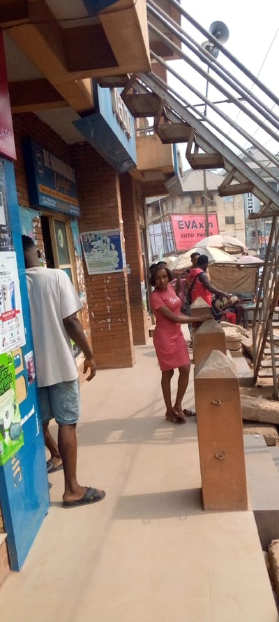 The image presents an outdoor urban commercial scene in Nnewi, Nigeria, during daylight.
In the foreground, a woman wearing a short-sleeved, knee-length red dress with "Barbie" printed in white, and a patterned headband, stands facing the camera with a slight smile. Her right hand rests on a chamfered brown concrete pillar, which features two metal rings embedded on its visible side. To her left, partially obscured by a blue vertical sign displaying mobile phone advertisements, a man in a light-colored t-shirt, denim shorts, and dark sandals stands with his back largely to the camera. The ground is a light-colored tiled or concrete surface, displaying shadows cast by figures and structures.
Behind the woman, a second identical brown concrete pillar is visible. To the right, an unpainted metal staircase with horizontal rails and zig-zag steps ascends towards the upper right of the frame. In the mid-ground, behind these structures, a market area operates under several light-colored fabric umbrellas supported by frames. Multiple individuals are visible within this market, including two men wearing red tops. Further into the background, a prominent red rectangular sign is mounted, displaying "EVAxon AUTO PARTS" and partial contact information.
The immediate background on the left shows a building facade made of brown brick. A blue sign above a doorway reads "DE HONORABLE Gadget," indicating "Dealers on all sorts of Phone, Phones & Accessories." A white poster depicting various electronic devices is affixed to this brick wall. Overhanging architectural elements with rectangular cutouts are visible above. High in the background, behind the market and staircase, two grey horn loudspeakers are mounted on a pole. Additional multi-story buildings are faintly visible in the distance under a bright sky.