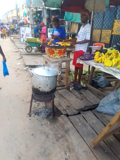 This is an outdoor street scene in Egbagu, Nigeria, likely a marketplace. A woman in a blue t-shirt and another woman in a checkered shirt and red skirt are standing at a food stall. The woman in blue is holding a phone. In front of them, corn is being cooked in a large metal pot over a charcoal stove. There are also bananas, what appear to be plums or dates, and corn husks visible at the stall. In the background, other people and motorcycles are present, along with various market stalls and buildings. The ground is a mixture of dirt and pavement.