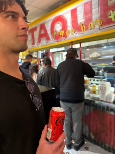 The image depicts the interior of Taquilla Taco Bar, located in Boston, United States. In the foreground, a male individual with dark, curly hair is seen in profile, looking towards the right side of the frame. He is wearing a black v-neck shirt, with a pair of dark-framed sunglasses hooked onto the collar. His right hand holds a red Coca-Cola Zero Sugar can, displaying a "RECYCLE ME" label.
In the midground, several other patrons are visible, standing in a line at a service counter. One man, seen from the back, wears a black puffer jacket and light grey pants, facing the counter. To his left, another man in a dark jacket is also facing the counter, and a woman with light brown hair is partially visible between them. Further left, another man in a dark hoodie is visible.
Above the counter, a large sign with a yellow border features the word "TAQUILLA" in prominent red letters, with "Taco bar" in smaller, yellow cursive letters below it. The serving counter below the sign displays various containers and a clear sneeze guard, suggesting food preparation or serving. The lower portion of the counter is constructed from corrugated metal, painted red. The floor is tiled with a white and brown geometric pattern.