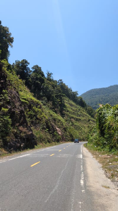 The image depicts a road winding through a mountainous region near the city of Unknown in Vietnam. The road surface is asphalt, with visible yellow lane markings. A dark-colored vehicle is present on the road. The landscape features steep hills covered in lush green vegetation. The sky is clear and blue, suggesting a sunny day. A section of the road has a dirt shoulder. The vegetation includes a variety of trees and smaller plants clinging to the rocky slopes.