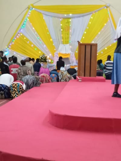 The image shows a large, brightly lit room with a stage in the foreground. The stage is covered in pink carpet and has a single step. The room is decorated with yellow and white curtains and a white cross hanging from the ceiling. There is a wooden lectern to the right of the stage and a group of people kneeling in prayer on the carpet in front of the stage. The people are mostly dressed in casual clothing, though some are wearing traditional African attire. The room is likely in the city of Arusha, Tanzania, based on the coordinates provided.  The image suggests that this is a religious event or service.  The people in the room are facing a white alter, with a white cloth draped over it, at the back of the room.  There are decorations in the background, likely flowers and a candle, but it is not clear what the exact decor is. There is also a person wearing a white head covering in the background, but they are out of focus.