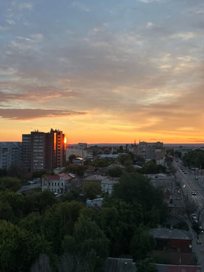 This aerial, high-angle shot captures a cityscape of Novokuybyshevsk, Russia, during a sunset. The sky is ablaze with vibrant hues of orange and yellow, with scattered clouds tinged with pink and blue. In the mid-ground, a tall, rectangular apartment building stands out, its glass facade reflecting the brilliant sunset. Adjacent to it are other buildings of varying heights and architectural styles, some appearing older with red-tiled roofs, others more modern. A tree-lined street runs through the city, with cars visible, indicating ongoing activity. Dense green foliage dominates the foreground, obscuring some of the lower structures and suggesting the presence of parks or residential gardens. The overall impression is a blend of urban development and natural elements, bathed in the warm glow of the setting sun.