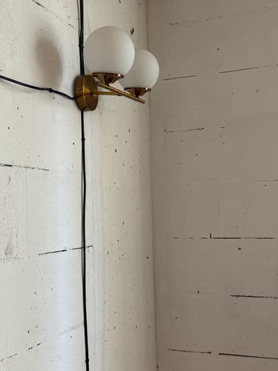 The image shows a close-up, high-angle shot of a brass double-globe wall sconce mounted on a textured, off-white concrete wall. The sconce has two frosted white spherical glass shades. Black electrical wire is attached to the sconce with plastic clips and runs vertically down the wall. The wall has horizontal lines and dark specks, suggesting it may be unfinished or aged concrete. The setting appears to be indoors, potentially in a residential or commercial space. There is no indication of the city of Irpin, Ukraine, or any specific location within it.
