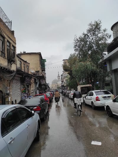 The image depicts a wet, narrow street or alleyway in what appears to be a Middle Eastern city, specifically Al-Qanawat Municipality, Syria. The scene is captured on an overcast day, indicated by the diffused light and the damp, reflective surface of the asphalt road, suggesting recent rainfall.

The street is lined on both sides with parked cars of various models and colors, including white, red, black, and silver/grey. A bicycle with a large, dark delivery bag on its rear rack is parked closer to the center of the street. Further down the street, a person wearing a light brown jacket walks away from the viewer, while another figure is visible near parked cars on the left.

On the left side of the street, older, multi-story buildings with weathered facades, some showing signs of disrepair like exposed brick and broken windows, line the street. Utility lines and cables crisscross above these structures. On the right, the buildings appear slightly more modern or better maintained. Above the rooftops in the distance, a prominent domed structure with a slender minaret, characteristic of a mosque, is clearly visible, providing a strong cultural and geographical clue. Lush green trees are interspersed among the buildings, particularly on the left.

The overall impression is one of everyday urban life in a bustling, historical area, under a quiet, rainy sky. No discernible text is visible on signs or vehicles.