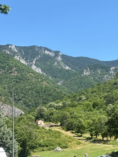 This image depicts a serene mountain landscape with a small stone church in Bachkovo, Bulgaria. The scene is dominated by lush green forested mountains under a clear blue sky. In the mid-ground, a small, traditional stone church with a red-tiled roof stands on a gentle slope surrounded by trees. A dirt path winds its way towards the church. A tall, modern streetlight is visible in the left foreground, along with a portion of a vehicle. In the far background, the rugged peaks of the mountains are visible, partially covered in vegetation and scree. The overall impression is one of natural beauty and tranquility.