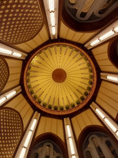 The image shows the interior of the Süleymaniye Mosque in Istanbul, Türkiye. The camera is pointed upwards towards the dome of the mosque. The dome is painted with a yellow and green pattern, with an inscription in Arabic script. The dome is illuminated by light fixtures that are mounted to the walls. The mosque's walls are painted a dark brown color and are covered in intricate geometric patterns. There is no one in the image.