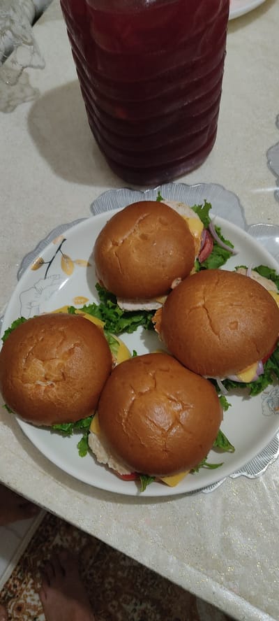 A plate of four mini burgers is presented on a table in Shamalgan, Kazakhstan. The burgers are arranged in a circular pattern on a white plate with a decorative rim. Each burger features a golden-brown bun, lettuce, cheese, and what appears to be thinly sliced meat and tomato. A dark red beverage in a ribbed container is visible in the background, partially obscuring the top of the plate. The table surface has a textured, light-colored finish, and a patterned rug is visible on the floor below. One person's feet are visible in the lower left corner of the image.