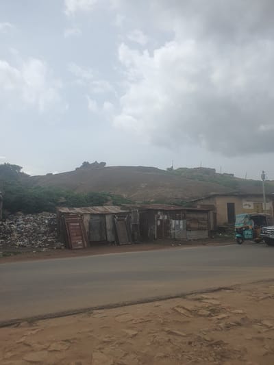 A wide-angle, medium-shot photograph captures a rural roadside scene with a rocky, vegetated hill in the background, located on Rukuba Road in Nigeria. The foreground features a paved road with a dirt shoulder. To the left, a large pile of refuse and debris is visible, alongside a cluster of makeshift, corrugated metal shacks. Further to the right, more shacks and a light-colored building with an arched doorway are present. A teal and white three-wheeled auto-rickshaw, identified by the license plate "PTA 2572," is parked on the right side of the road, next to a black vehicle. The sky is overcast with a mix of white and gray clouds.