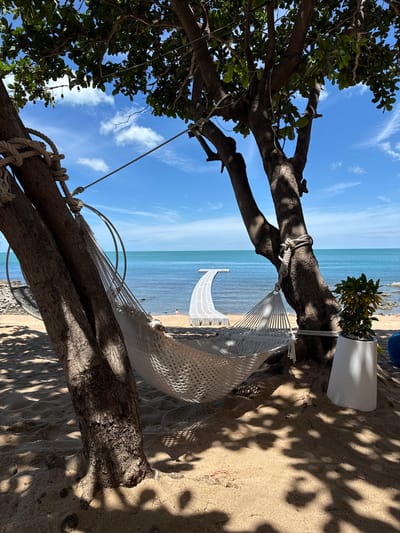 A white macrame hammock is suspended between two trees on a sandy beach in Choeng Mon, Thailand. The hammock is partially in shadow from the trees, which have green leaves and are illuminated by sunlight. The ocean is visible behind the hammock, with a clear blue sky above and some clouds. A white modular walkway extends into the water from the beach. In the foreground, the sand is textured with shadows cast by the trees. To the right of the trees, a white planter holds a green plant. A person can be seen in the ocean in the distance, and the rocky shoreline is visible to the right. The scene conveys a relaxed, tropical atmosphere.