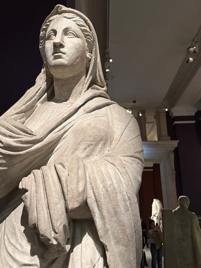 The image captures a close-up of a classical stone statue, likely a female figure, with detailed drapery and a serene facial expression. The statue is made of a light-colored material, possibly marble or limestone, and exhibits signs of age and wear. It is displayed indoors, as indicated by the ceiling with lighting fixtures and architectural elements in the background. The lighting is dramatic, casting shadows that highlight the contours of the statue. In the mid-ground, other statues and visitors can be observed, suggesting the setting is a museum or historical exhibition space. The overall ambiance is one of reverence and historical preservation. This statue is located at the Istanbul Archaeology Museums, which is situated in the Sultanahmet district of Istanbul, Turkey.