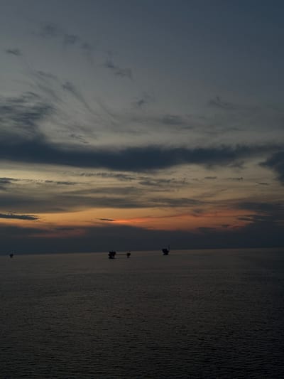 The image depicts a vast expanse of dark, rippling water under a twilight sky. The sky transitions from a deep, dusky blue at the top to lighter shades of gray and orange near the horizon, with wispy clouds scattered throughout. In the distance, silhouetted against the horizon, are several offshore structures, likely oil rigs or platforms. One structure, to the right of center, appears larger and more complex, connected by a walkway to another platform. Two smaller, simpler structures are visible further to the left, one closer and one more distant. The overall impression is one of a remote, industrial seascape at the end of the day, with the structures standing as solitary figures against the fading light. The location is identified as Unknown, Italy, but no specific urban context is discernible.