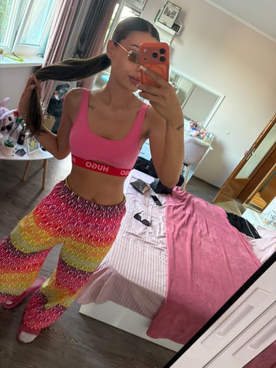 A woman is taking a selfie in a bedroom. She is wearing a pink sports bra with the "HUGO" logo on the band and colorful, striped pants. She has her hair in a ponytail and is wearing sunglasses. A large pink blanket is draped over a bed in the background. The room appears to be well-lit, suggesting it is daytime. The presence of a bed and a closet with mirrored doors indicates it's a bedroom. A phone is being used to take the photo, and the reflection in the mirror shows more of the room, including a dresser and a window with blinds. The overall mood seems casual and relaxed. There is text visible on the woman's sports bra ("HUGO") and a tattoo on her arm, though the text of the tattoo is not clearly legible. The location is identified as Karnobat, Bulgaria.
