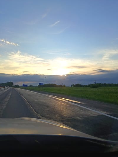 The image depicts a view from inside a vehicle driving on a highway near Malyi Sundyr', Russia. The road surface appears wet, reflecting the light of the setting sun. A grassy field borders the right side of the road, with a treeline visible in the distance. Signage is present on both sides of the road. The sky is a mix of blue and cloudy hues, with the sun low on the horizon. The vehicle's dashboard and hood are visible at the bottom of the frame.