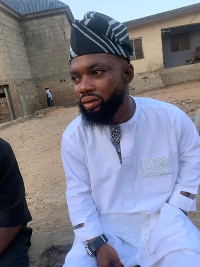 The scene is set outdoors in Okene, Nigeria, depicting a daylight shot of a male subject in the foreground, with an urban or semi-urban environment in the background.
The primary subject is a male, estimated to be in his late 20s to early 30s, positioned slightly off-center to the right, looking towards his right with a neutral expression. He has short, dark hair with faded sides, a full, neatly trimmed dark beard and mustache. He is wearing a white long-sleeved tunic (buba) with a round neck and an intricate, dark-patterned fabric visible at the inner collar opening. The tunic features a rectangular embellishment of small, light-colored studs or rhinestones on the left chest pocket area, forming a stylized design. A similar embellishment is visible on the lower front of the tunic, near the waist. He wears a black and white striped cap (fila) commonly worn in Yoruba culture. His left arm is visible, wearing a dark-strapped wristwatch with a silver-colored casing. His right hand is partially obscured, possibly in his pocket. A dark-clad individual is partially visible on the far left foreground.
The background consists of unpaved, sandy-colored ground. Behind the primary subject, there are multiple structures made of concrete blocks. To the left, a two-story building constructed from rough, unpainted concrete blocks with a dark, sloped roof is visible. A dark rectangular opening, possibly a doorway, is present on its lower level. Further back and to the left of this building, a smaller, slender figure is visible walking, carrying a reddish-brown object on their head. To the right of the central subject, another single-story building, possibly painted a light yellow or beige, with darker window openings, is visible. A seated figure is faintly discernible near this building. The sky is clear and light, suggesting daytime.