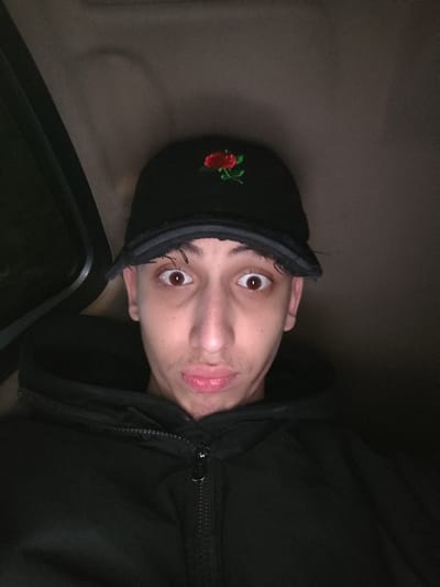 A close-up selfie captures a young male inside what appears to be the dimly lit interior of a vehicle. He has light skin, prominent dark brown eyes, and a light mustache and chin stubble. He is wearing a black baseball cap featuring a red rose embroidery on the front, and a black hooded zipper jacket. His eyes are wide open, and his lips are puckered in a distinct, possibly playful or surprised, expression.
The lighting is low, with the subject's face well-lit, suggesting the use of a flash or an internal light source. On the left side of the frame, a dark, indistinct structure, possibly a window frame or door panel of the vehicle, is visible. The background is mostly dark, with subtle variations in tone consistent with an enclosed space. There are no identifiable location cues within the image itself beyond the vehicle interior, though the provided context places the image in As-Salihiya Municipality, Syria. No visible text is present in the image.
