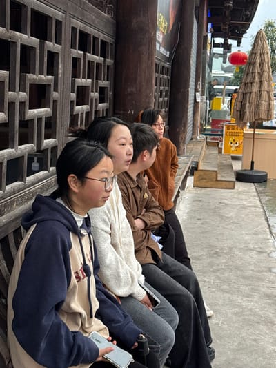 Four individuals are seated on a wooden bench against a traditional dark wooden building facade in Pengshui Miao and Tujia Autonomous County, China. From left to right: a female wearing glasses and a navy and beige hooded sweatshirt, holding a smartphone in a clear case; a female in a light-colored knitted sweater; a male in a brown collared shirt with arms crossed; and a female wearing glasses and a brown fuzzy jacket, leaning back. All individuals appear to be looking to their right, off-frame. The building features dark wooden lattice panels and vertical wooden pillars. The ground in front of the bench is a wet, grey paved surface. In the background, a red Chinese lantern is visible, along with a conical thatched parasol on a black base, and a yellow sign featuring a cartoon tiger. The overall environment suggests an urban or town setting, with overcast weather.