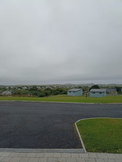 The image depicts a scenic view of Doirí Beaga, Ireland, under a heavy overcast sky. In the foreground, a dark asphalt driveway meets a neatly mowed green lawn bordered by a grey concrete curb. The driveway leads to a paved walkway with rectangular grey stones.

Beyond the lawn, a wooden fence encloses a grassy area with playground equipment, including a swing set and a wooden climbing structure. Further in the background, a small village is visible, characterized by numerous white houses with grey roofs, interspersed with trees and green hills. Two light blue, modern-style sheds with dark grey roofs are situated on the grassy slope in the middle ground. The overall atmosphere is tranquil and somewhat muted due to the cloudy weather.