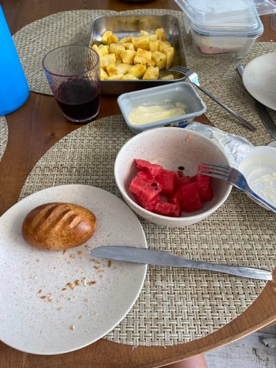 A wooden table, covered with a woven placemat, is set with a variety of food items. There is a single bread roll on a white plate, a bowl of cubed watermelon, a glass of red wine, a container of what appears to be butter, and a metal dish with cubed pineapple. In the background, a clear plastic container with a plastic lid is visible. The scene appears to be a casual, home setting. The scene was captured in Punta Hermosa, Peru, at latitude -12.326, longitude -76.834.
