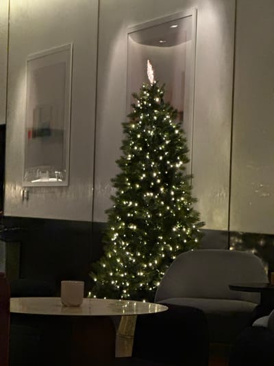 A lit Christmas tree is positioned against a light gray wall, partially visible through a large rectangular opening. An additional rectangular frame is below. A modern-style chair with a curved back is situated nearby, adding to the interior decor. An abstract sculpture or object with reflective surfaces stands at the front. The overall ambiance suggests a contemporary, holiday-decorated space.