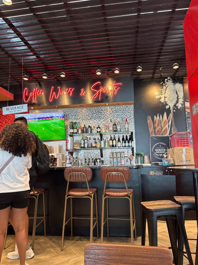 This is a waist-up, eye-level, medium shot of a bar area at "Coffee Wines & Spirits" in Mai Khao, Thailand. A neon sign in cursive red letters spells out "Coffee Wines & Spirits" above a black counter and a mosaic tiled backsplash. A large television screen displays a soccer match. On the shelves behind the bar are numerous bottles of alcohol and cups. Two bar stools with brown leather seats and gold metal frames are visible. In the foreground, a person with dark curly hair wearing a white t-shirt and black shorts stands with their back to the camera. Another person, only their legs are visible, stands next to them wearing white sneakers and black shorts. The flooring is made of light-colored wood planks. To the right of the bar, a black wall features a large graphic of bread loaves and flour. Red and white crates are stacked near the wall.
