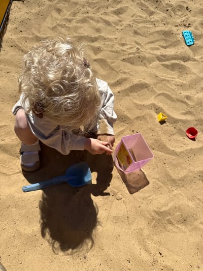 In a sandbox filled with light brown sand, a toddler with curly blonde hair is playing. The toddler is wearing a light grey long-sleeved shirt and light-colored shoes. They are in a crouching position, with their hands in the sand. In front of the toddler is a blue plastic shovel. To the toddler's right, there is a translucent pink plastic container, a small yellow triangular toy, and a red plastic cup. In the background, further to the right, a blue toy brick can be seen. The scene is illuminated by bright sunlight, casting shadows on the sand. The location is a sandbox, possibly in a playground, and it is not possible to determine the specific location within the city of Vladimir, Russia, from the image alone.