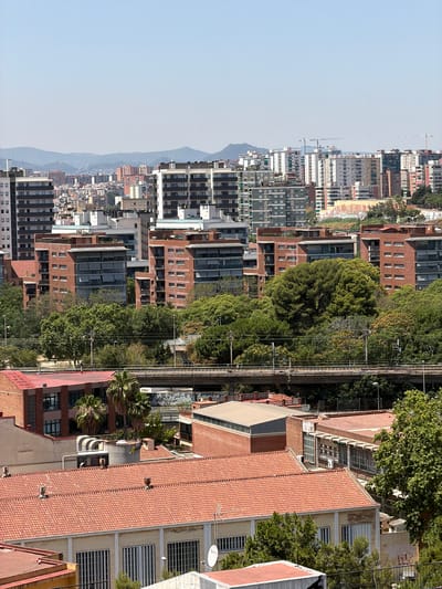 This is an aerial, rotated image of a densely populated urban area with a clear sky, suggesting daytime. The scene depicts a cityscape with numerous multi-story buildings, some appearing residential and others more commercial or industrial, characterized by brick facades and various window designs. Trees, some large and mature, are interspersed throughout the urban landscape, adding a touch of greenery. A prominent feature is a set of railway tracks running vertically through the image, with overhead power lines and supporting structures. In the background, a hazy mountain range can be seen, and further in the distance, several construction cranes are visible, indicating ongoing development. The architecture suggests a European setting, consistent with the provided location context of l'Hospitalet de Llobregat, Spain. There are no people clearly visible in the image, and the overall mood is one of a bustling, active city. Some graffiti is visible on a building in the upper right quadrant, and what appears to be a satellite dish is mounted on one of the buildings.