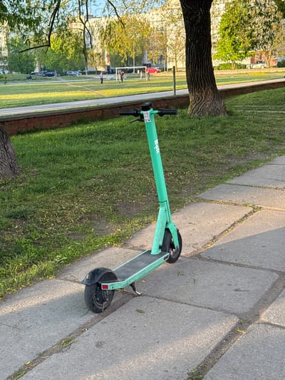 An electric scooter, branded "Bolt," is parked on a gray concrete sidewalk adjacent to a grassy area. The scooter is light green. The grass area is bordered by a red brick wall and a track, suggesting a park or recreational field nearby. In the background, there is a partially obscured view of buildings and trees, characteristic of an urban environment. People are visible on the track and field. Based on the architecture, general environment, and the Bolt scooter branding, the location is likely a park in Liepaja, Latvia.