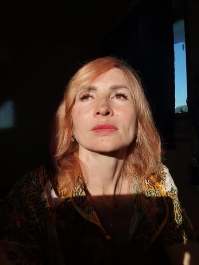 A close-up shot captures a woman with auburn hair looking upwards and to the left, illuminated by a strong light source from the right. Her face is partially in shadow, creating a dramatic effect. She wears a brightly patterned shirt with a tiger print and floral accents. A silver necklace with a small pendant is visible. The background is dark, with a sliver of bright blue light visible on the right, suggesting a window or opening. There are no other people or animals present. The scene appears to be indoors, possibly in a residential setting within the city of Bol'shie Mkhi, Russia, given the context. The lighting suggests it is either early morning or late afternoon.