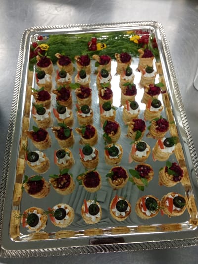 This image captures a large, ornate silver serving tray filled with a meticulously arranged assortment of appetizers, likely prepared for a catering event or a formal occasion. The rectangular tray features a decorative, ridged border and a highly reflective surface, mirroring elements of the surrounding environment, such as overhead lighting.
The tray holds numerous individual puff pastry canapés, arranged in neat rows. There are two distinct types of fillings, alternated for visual appeal. One variety features a creamy white topping, possibly a cheese or savory mousse, garnished with a slice of black olive and often a small sprig of fresh dill, sometimes accompanied by a thin strip of red bell pepper. The second type contains a rich, dark red, granular filling, strongly suggesting a beetroot-based mixture. These are typically adorned with a half-walnut and a small green leaf, possibly basil or mint.
At the top edge of the tray, fresh green dill sprigs, clusters of bright red berries (resembling redcurrants), and a few small yellow decorative pieces are used as garnishes, adding color and freshness to the presentation.
The tray rests on a smooth, metallic surface, likely a stainless steel countertop, indicative of a professional kitchen or catering preparation area. The lighting is bright and even, suggesting an indoor setting. No people or visible text are present in the image. The presentation highlights careful preparation and fresh ingredients.