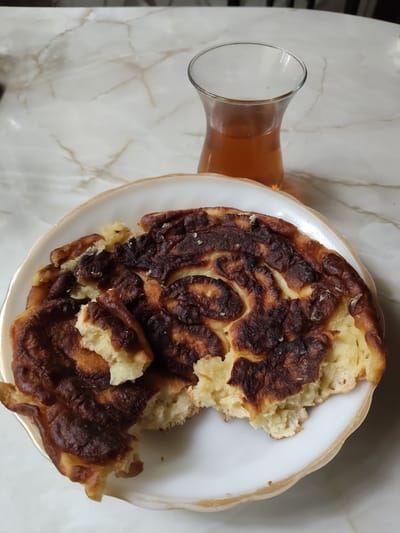 A plate of partially eaten pancakes and a glass of tea are on a marble table. The pancakes are golden brown with some burnt edges, and one has a bite taken out of it. The tea is a light brown color. The table is white with gray veining. The scene is lit from above. There is no indication of the city of Венюково, Russia.