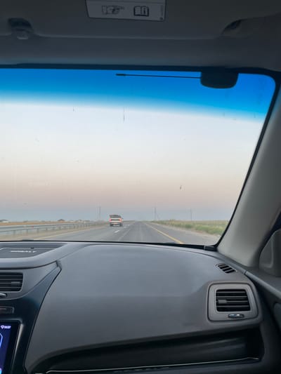 The image is taken from inside a car, looking out through the windshield. The dashboard of the car is visible in the foreground. The road ahead is straight and appears to be a highway. There is a white pickup truck driving in the same direction as the camera car. The sky is a pale gradient of blue and pink, suggesting it might be dawn or dusk. The environment appears to be rural or on the outskirts of a town, with sparse vegetation and a flat landscape. There are no people clearly visible in the image besides the driver implied by the camera's perspective. The car appears to be in motion, driving on a paved road. The location is specified as Toksanbay, Kazakhstan.